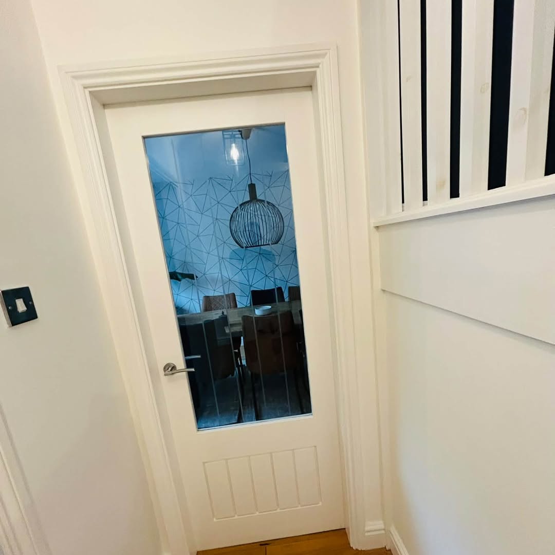 A white painted panelled wall beside a glazed internal door with black painted spindles behind.